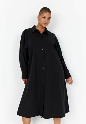 Femeie cu păr scurt purtând o rochie midi neagră cu mâneci lungi, nasturi și croială lejeră, stând în picioare pe un fundal alb.