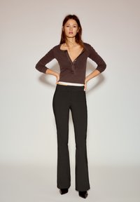 Tamno smeđa majica dugih rukava s gumbima na izrezu, uparena s crnim zvonolikim hlačama, obje odjeće uske i glatke teksture.