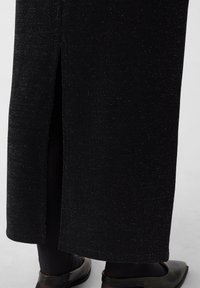 Czarna spódnica ołówkowa z delikatnym połyskiem, z bocznym rozcięciem. Wykonana z teksturowanej tkaniny. Noszona z czarnymi butami.