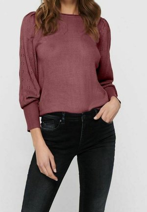 Femme portant un haut en maille mauve à manches longues avec manches bouffantes en dentelle et un jean noir taille haute, une main dans la poche.
