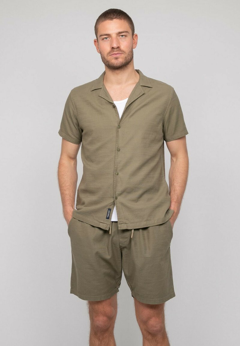 Chemise à manches courtes vert olive clair avec un devant boutonné et une coupe décontractée, assortie à un short. Le tissu semble doux et texturé.