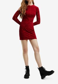 Femme en robe mini moulante à manches longues rouge à motifs, portant des bottines noires à lacets, debout avec les mains derrière le dos.