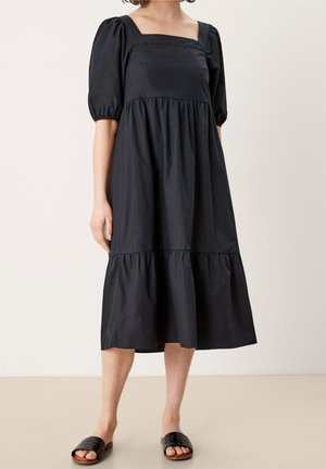 Kobieta ubrana w czarną sukienkę o długości midi z bufkami na łokciach, kwadratowym dekoltem, marszczoną talią i czarnymi plecionymi klapkami.