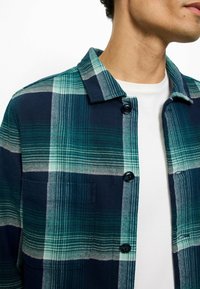 Flanellhemd in blau-grünem Kariertes Muster, mit einem Kragen mit Knöpfen, zwei Knöpfen und einer Brusttasche. Baumwollstoff mit weicher Textur.