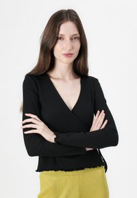 Černý žebrovaný top s dlouhým rukávem a hlubokým výstřihem do V, s volánkovým lemem a manžetami, spárovaný se žlutými texturovanými kalhotami.