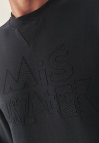 Czarna bluza z podniesionymi, teksturowanymi literami w kontrastującym, czarnym wzorze. Żebrowany dekolt i charakterystyczny trójkątny szew.