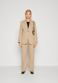 Costum din in bej, cu o jachetă ajustată și pantaloni largi. Top negru dedesubt, asortat cu pantofi negri fără toc și un brad dintr-o curea argintie.