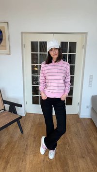 Moteris stovinti viduje su kišenėse rankomis, dėvi baltą kepurę, rožinę dryžuotą megztinį, juodas kelnes ir baltus sportbačius su rožiniais akcentais.