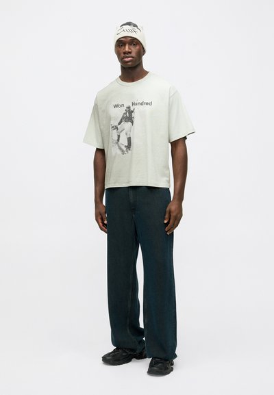 Λαμπερό πράσινο oversized t-shirt με γραφικό τύπωμα και κείμενο, συνδυασμένο με σκούρα μπλε φαρδιά παντελόνια και μαύρα υποδήματα. Απλό πλεκτό καπέλο.