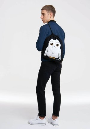 Schwarzer Kordelzug-Rucksack aus Stoff, mit einem weißen Eulen-Motiv mit gelben Augen und orangen Füßen, glatte Oberfläche und verstellbaren Trägern.