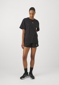 Black cotton T-shirt with a crew neckline and short sleeves, paired with black shorts. White logo accents on both items; model wearing black sneakers.