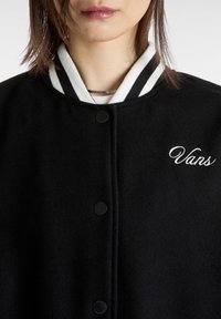 Svart ull varsityjacka med vit och svart randig ribbad krage, som har svarta tryckknappar och en vit broderad "Vans"-logotyp.
