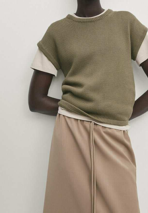 Personne portant un haut sans manches en maille vert olive sur une chemise beige à manches courtes et une jupe beige avec une couture à l’avant.