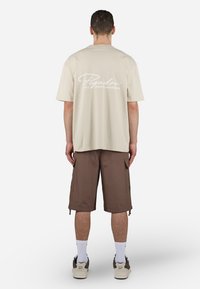 Beige oversized t-skjorte med hvit "Pegador"-logo på baksiden, sammen med brune cargo-shorts. Modellen har på seg hvite sokker og brune sneakers.