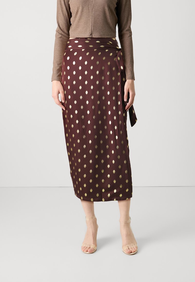 Femme portant une jupe portefeuille midi marron à pois dorés, des sandales beiges à talons hauts et un top marron côtelé à manches longues.