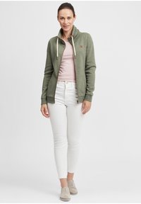 Grønn zip-up jakke med høy krage, kremfargede snorer, båret over en lys rosa topp, sammen med hvite skinny jeans og lyse sko.