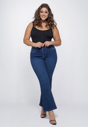 Vrouw met golvend haar lacht, draagt een zwarte mouwloze top, blauwe wijde jeans en zwarte sandaalhakken, staand tegen een effen achtergrond.