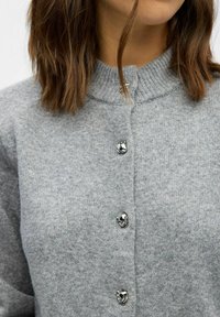 Grijze gebreide cardigan met een hoge kraag, vijf metalen knopen en een zachte textuur. Het kledingstuk heeft een eenvoudig, strak ontwerp.