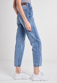 Mujer con pantalones vaqueros de cintura alta y zapatillas blancas, de pie de lado contra un fondo claro y liso.