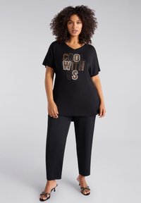 Camiseta negra de manga corta con la inscripción "COOL WITH US" en tachuelas doradas y plateadas, combinada con pantalones negros y tacones negros con tiras.