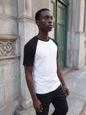 Νέος άνδρας με λευκό και μαύρο T-shirt στέκεται κοντά σε ένα λιθόκτιστο κτήριο με περίτεχνα σκαλίσματα και μια πράσινη πόρτα.