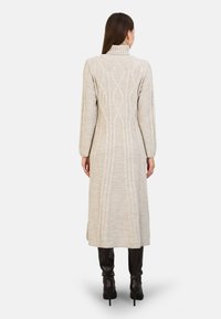 Robe tricotée beige longue avec un col montant, présentant un motif en câble texturé au dos et une coupe droite, atteignant la longueur mi-mollet.