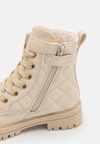 Botín de tobillo beige con cordones, patrón acolchado, ojales en tono dorado y cremallera lateral. Suela de goma y forro suave para mayor comodidad.