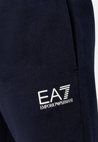 Pantaloni jogging navy realizzati in tessuto morbido, con tasca laterale e logo bianco EA7 Emporio Armani sulla coscia. Texture liscia e design casual.