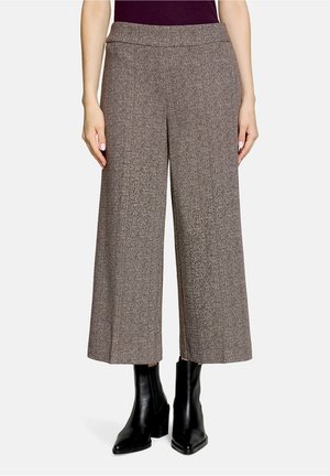Kvinde iført højttaljede, viddebrune cropped tweedbukser og sorte ankelstøvler, stående mod en ensfarvet hvid baggrund.