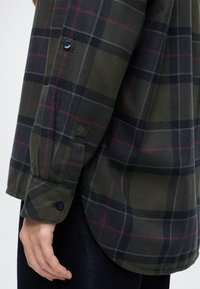 Gros plan d'une manche de chemise à carreaux vert foncé et noir avec un poignet boutonné, portée par une personne dont la main est détendue à côté de son pantalon foncé.