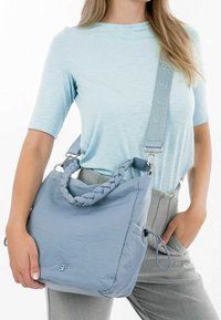 Sac bleu clair avec une poignée tressée et une bandoulière réglable, présentant une texture lisse et un logo subtil à l'avant.