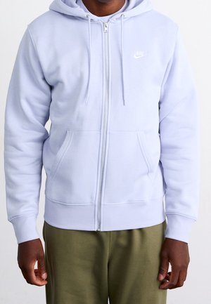Personne portant un sweat à capuche zippé Nike violet clair et un pantalon vert olive sur un fond uni.