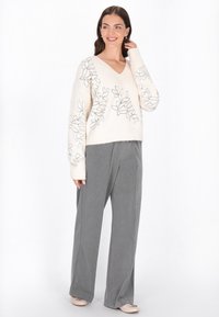 Kremowy sweter z dekoltem w serek z czarnym haftem liści, w zestawieniu z luźnymi szarymi spodniami. Gładka tekstura, długie rękawy i luźny krój.
