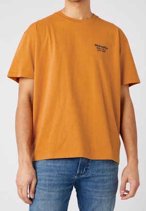 Homme portant un t-shirt orange à manches courtes Wrangler avec un texte sur la poitrine et un jean bleu, montré du cou aux hanches.