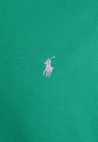 Grön bomullströja med en liten lila broderad logotyp av en polo-spelare som rider på en häst. Slät textur, rund halsdesign.