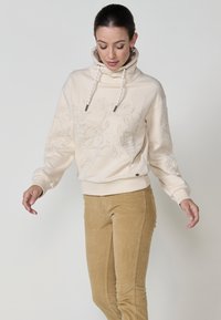 Sweat à capuche crème avec broderies florales, col montant et cordons. Associé à un pantalon en velours côtelé marron clair avec une poche latérale.