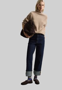 Maglione a coste beige, jeans a vita alta blu scuro con orli arrotolati, mocassini marroni e una borsa a tracolla marrone tenuta in una mano.