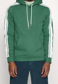 Sudadera verde con rayas blancas en los laterales, capucha con cordones y bolsillo frontal tipo canguro. El tejido parece suave, probablemente una mezcla de algodón, con puños acanalados.