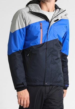 Homme portant une veste à blocs de couleurs bleu, noir et gris avec une capuche et des poches zippées, debout devant un fond uni.