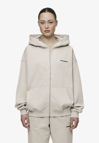 Bežna zip-up dukserica napravljena od mekog materijala, sa velikom kapuljačom, spuštenim ramenima i prednjim dzepovima. Crni logo na lijevoj strani grudi.