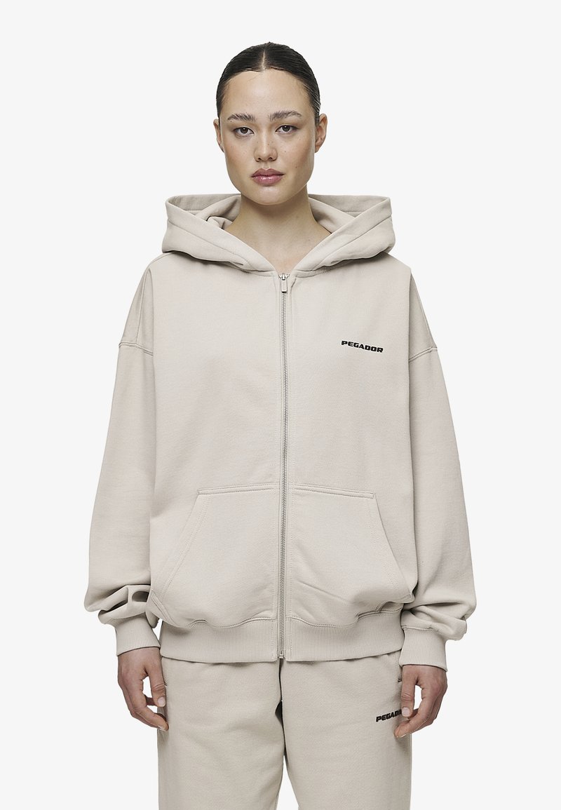 Bežna zip-up dukserica napravljena od mekog materijala, sa velikom kapuljačom, spuštenim ramenima i prednjim dzepovima. Crni logo na lijevoj strani grudi.