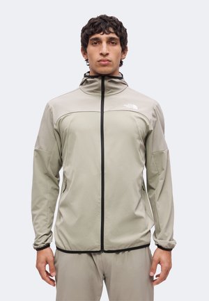 Mand med kort krøllet hår iført en lysebeige The North Face jakke med lynlås foran og stående foran en ensfarvet baggrund.