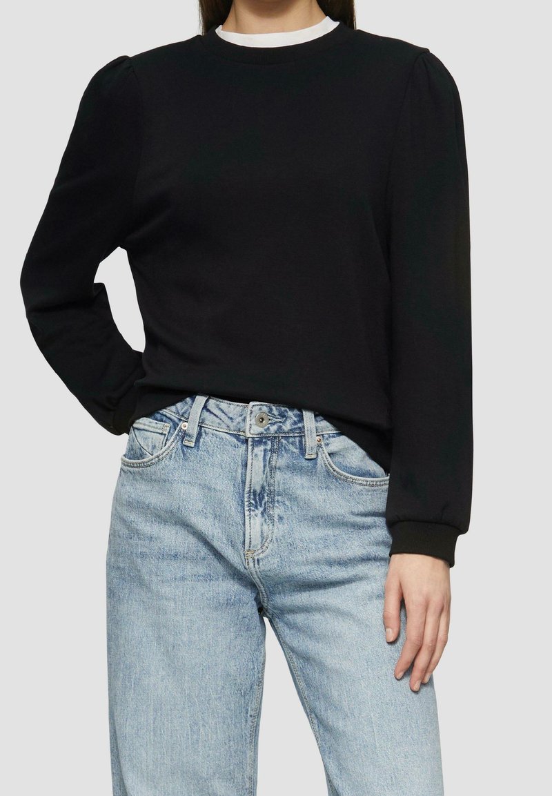 Femme portant un pull noir à manches longues avec un col blanc et un jean taille haute bleu clair, une main sur la hanche et l'autre détendue.
