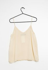 Fließende beige Spaghetti-Tops mit dünnen Trägern, V-Ausschnitt und softem Material. Hängt an einem schwarzen Haken vor einem schlichten Hintergrund. Transparente Etikette sichtbar.