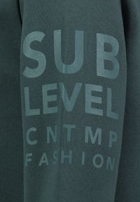 Groene sweatshirt met een zachte textuur, voorzien van grote, gedrukte tekst: "SUB LEVEL CNTMP FASHION" in een contrasterende lichtere tint.