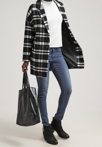 Cappotto a quadri bianco e nero con colletto, indossato sopra una maglia bianca, abbinato a jeans blu e stivali neri alla caviglia. Borsa tote nera in mano.