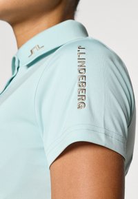 Ljusblå pikétröja med krage, korta ärmar och broderad text "J. Lindeberg" på vänster axel. Mjuk tygstruktur.