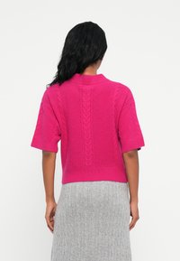 Pull court à manches courtes de couleur rose vif, avec une texture en maille torsadée et un ourlet côtelé. Associé à une jupe côtelée gris clair.