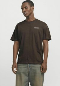 Brown cotton t-shirt with short sleeves, featuring a small "FIREPLACE" logo in white on the left chest. Relaxed fit and crew neck design.