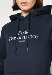 Felpa con cappuccio blu navy realizzata in cotone misto, con logo ricamato bianco "Peak Performance" e "Est. 1986" sul davanti.